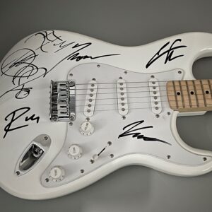 A white electric guitar with a maple neck and multiple signatures on the body. The guitar is viewed from the side, highlighting its pickups, controls, and the signed surface.