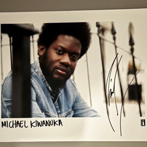 A portrait of Michael Kiwanuka wearing a denim jacket, sitting on a staircase. The image features a white border with the name 'MICHAEL KIWANUKA' written below, along with an autograph.